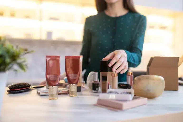 A woman in a green blouse organizes assorted beauty products, including tubes, bottles, and brushes on a white table. The setting appears modern and well-lit.