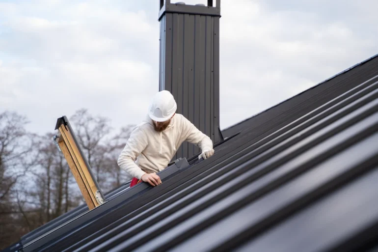 A construction worker in a white hard hat inspects a modern metal roof, with a chimney nearby. The sky is overcast, suggesting a calm atmosphere.