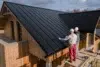 Two workers in helmets install a sleek black metal roof on a modern wooden house under a partly cloudy sky, conveying teamwork and efficiency.