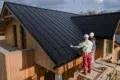 Two workers in helmets install a sleek black metal roof on a modern wooden house under a partly cloudy sky, conveying teamwork and efficiency.