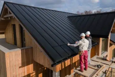 Two workers in helmets install a sleek black metal roof on a modern wooden house under a partly cloudy sky, conveying teamwork and efficiency.