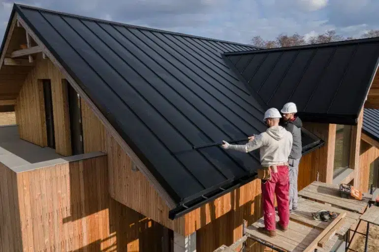 Two workers in helmets install a sleek black metal roof on a modern wooden house under a partly cloudy sky, conveying teamwork and efficiency.