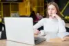 Person in a white sweater sitting at a table with a laptop, holding a cup of coffee and an open book in a cafe.