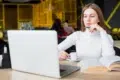 Person in a white sweater sitting at a table with a laptop, holding a cup of coffee and an open book in a cafe.