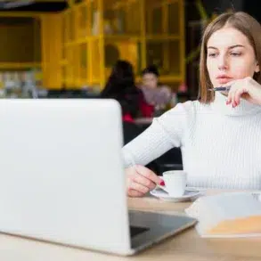 Person in a white sweater sitting at a table with a laptop, holding a cup of coffee and an open book in a cafe.