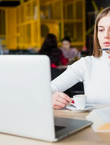 Person in a white sweater sitting at a table with a laptop, holding a cup of coffee and an open book in a cafe.