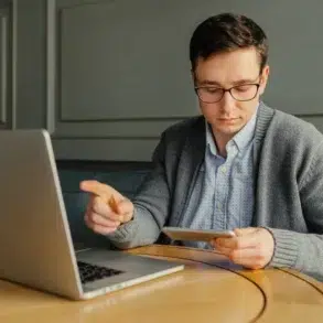 A man in glasses sits at a round wooden table, focused on a tablet, with a laptop open nearby. The setting is calm and studious.