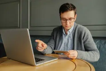 A man in glasses sits at a round wooden table, focused on a tablet, with a laptop open nearby. The setting is calm and studious.