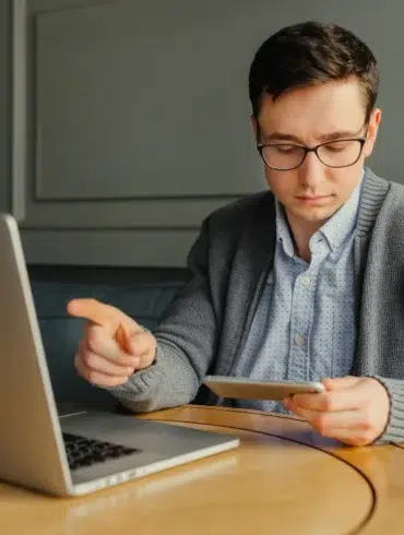 A man in glasses sits at a round wooden table, focused on a tablet, with a laptop open nearby. The setting is calm and studious.