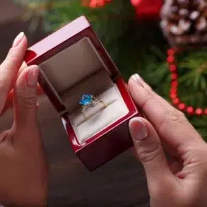 Hands hold an open red ring box with a blue gemstone ring inside. The background features festive decor, including pine branches and red ornaments, conveying a celebratory tone.