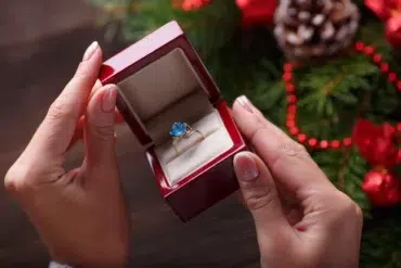 Hands hold an open red ring box with a blue gemstone ring inside. The background features festive decor, including pine branches and red ornaments, conveying a celebratory tone.