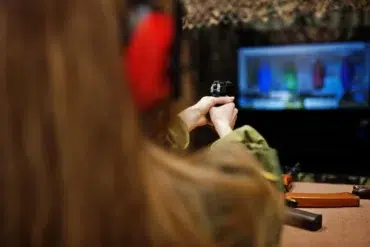 A person in red earmuffs aims a handgun at a digital shooting range screen, displaying colored targets. The focus is on concentration and precision.