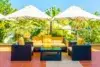 Outdoor patio with dark wicker furniture, yellow cushions, and striped pillows. Two large white umbrellas provide shade. Lush greenery in the background.