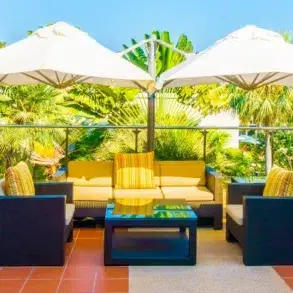 Outdoor patio with dark wicker furniture, yellow cushions, and striped pillows. Two large white umbrellas provide shade. Lush greenery in the background.