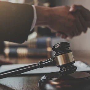 Wooden judge's gavel on table with two people shaking hands in the background, symbolizing legal agreement or settlement.