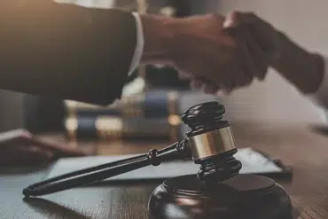 Wooden judge's gavel on table with two people shaking hands in the background, symbolizing legal agreement or settlement.