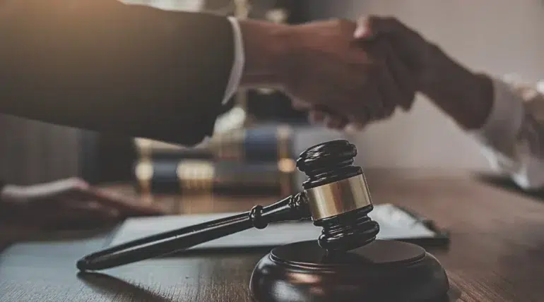 Wooden judge's gavel on table with two people shaking hands in the background, symbolizing legal agreement or settlement.