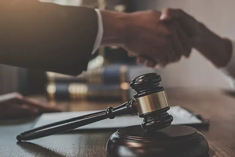 Wooden judge's gavel on table with two people shaking hands in the background, symbolizing legal agreement or settlement.