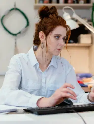 Woman with red hair in a white shirt working on a computer at a desk in a workshop with tools hanging on the wall behind her.