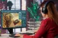 A woman wearing headphones works at a computer with a 3D modeling software displaying a room layout. The setting is a modern office with green plants.