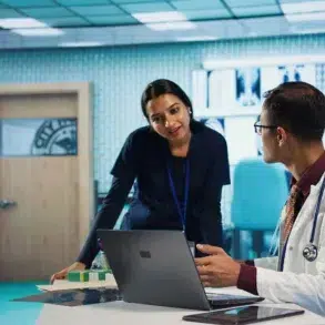 A doctor and a nurse converse in a hospital setting, with medical images displayed on a wall. The atmosphere is professional and collaborative.