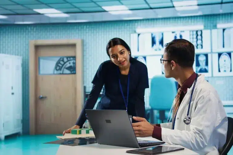 A doctor and a nurse converse in a hospital setting, with medical images displayed on a wall. The atmosphere is professional and collaborative.