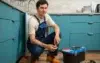 Plumber in blue overalls squatting by open kitchen cabinet, holding a pipe fitting with a toolbox and tools on the floor nearby.