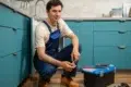 Plumber in blue overalls squatting by open kitchen cabinet, holding a pipe fitting with a toolbox and tools on the floor nearby.