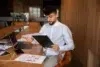 A man in a white shirt reviews documents at a wooden table, surrounded by charts and a laptop. The office setting and his focused expression suggest analysis.