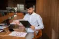 A man in a white shirt reviews documents at a wooden table, surrounded by charts and a laptop. The office setting and his focused expression suggest analysis.
