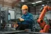 A worker in a blue uniform and yellow hard hat operates machinery in a factory. He is focused, using a control panel near an orange robotic arm.
