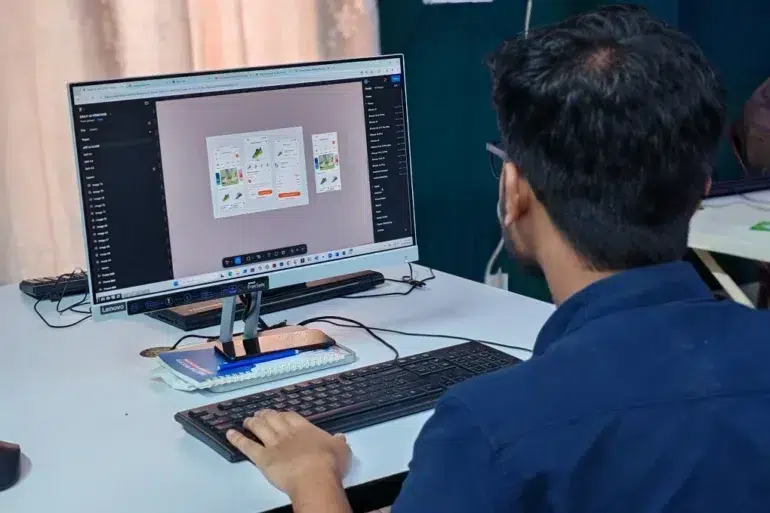 A person wearing glasses is focused on designing a mobile app interface on a computer monitor. The desk is cluttered with a keypad and tech accessories.