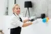 A woman in a white shirt and black apron dusts a black Buddha statue with a colorful feather duster, smiling, creating a clean, serene atmosphere.
