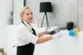 A woman in a white shirt and black apron dusts a black Buddha statue with a colorful feather duster, smiling, creating a clean, serene atmosphere.