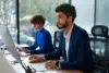 Two professionals in an office setting, a man in a blue suit and a woman in a blue sweater, work on computers by a large window.