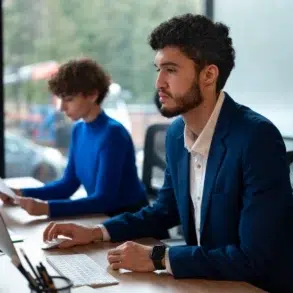 Two professionals in an office setting, a man in a blue suit and a woman in a blue sweater, work on computers by a large window.