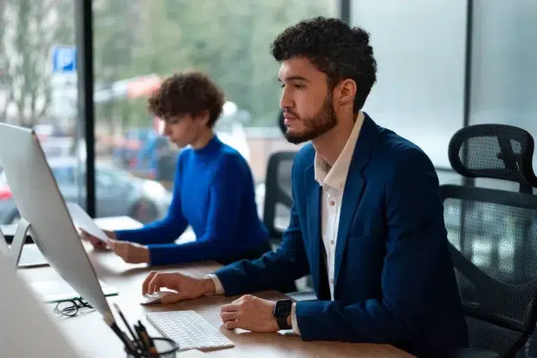 Two professionals in an office setting, a man in a blue suit and a woman in a blue sweater, work on computers by a large window.