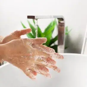 Person washing hands with soap under running water at a modern sink with a green plant in the background.