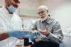 A dentist in gloves and mask shows a dental model to an elderly man in plaid shirt, sharing a lighthearted moment in a dental office setting.