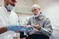 A dentist in gloves and mask shows a dental model to an elderly man in plaid shirt, sharing a lighthearted moment in a dental office setting.