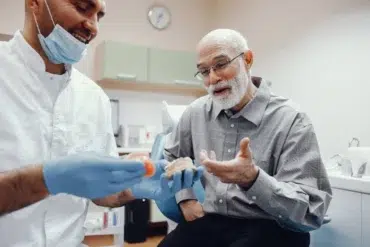 A dentist in gloves and mask shows a dental model to an elderly man in plaid shirt, sharing a lighthearted moment in a dental office setting.