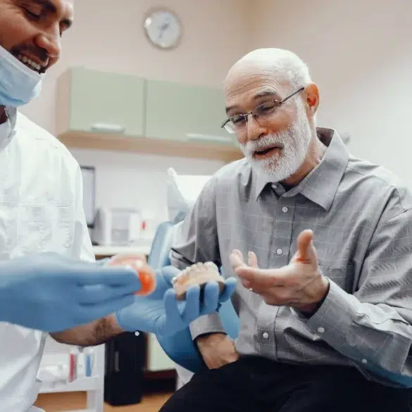 A dentist in gloves and mask shows a dental model to an elderly man in plaid shirt, sharing a lighthearted moment in a dental office setting.