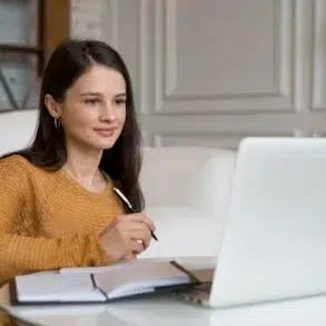 A young woman in a yellow sweater holds a pen while looking at a laptop and notebook.