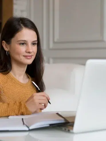 A young woman in a yellow sweater holds a pen while looking at a laptop and notebook.