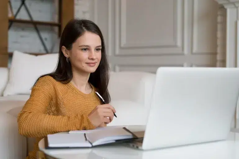 A young woman in a yellow sweater holds a pen while looking at a laptop and notebook.