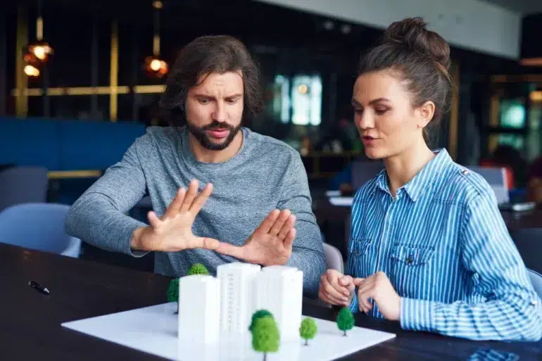 A man and woman are focused on a white architectural model with trees on a table. The man gestures while discussing the model. The setting is modern and collaborative.