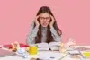 Student with red-rimmed glasses holding head in hands with open book and crumpled papers on desk.