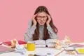 Student with red-rimmed glasses holding head in hands with open book and crumpled papers on desk.
