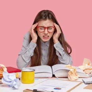 Student with red-rimmed glasses holding head in hands with open book and crumpled papers on desk.