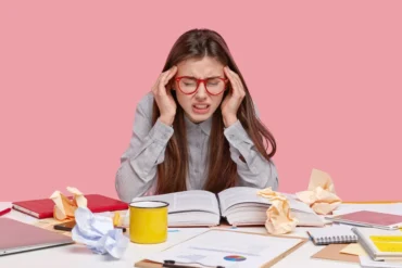 Student with red-rimmed glasses holding head in hands with open book and crumpled papers on desk.